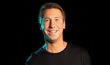 Close-up photo of Carl Hutchinson smiling at the camera, wearing a black t-shirt on a black background. There is a blue light shining on one side of his face