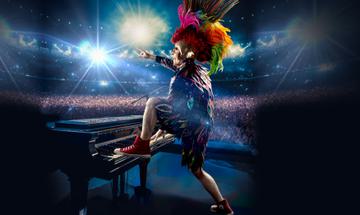 A tribute performer dressed as Elton John (A colourful jacket and head piece) with his foot on a piano looking out to the audience