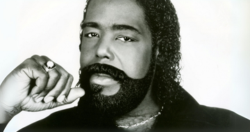 A black man with long curly hair and a thick beard holds his hand to his face