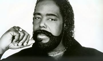 A black man with long curly hair and a thick beard holds his hand to his face