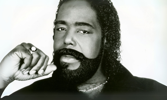 A black man with long curly hair and a thick beard holds his hand to his face