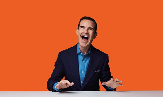 Jimmy Carr sat behind a desk with his hands slightly raised and his mouth open, wearing a suit. He is in front of a bright orange background