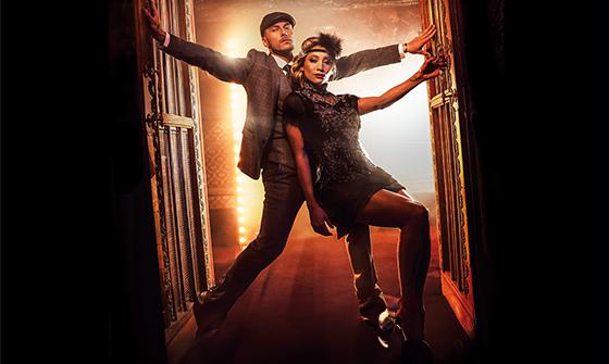 Gorka Marquez and Karen Hauer stood  in a doorway with red/orange light behind them and black in front. Gorka is in a three piece grey suit with a flat cap holding the door frame. Karen us in a flapper outfit leaning on Gorka holding the right side of the door