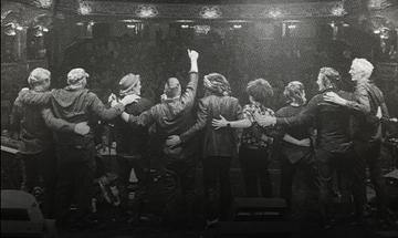 Black and white photo of the backs of nine members of the Levellers on stage, looking towards the audience with their arms around each other