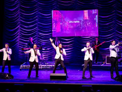 Five men on stage in front of a band behind with a keyboard, guitar, bass, and drum set. The five men are in white suit jackets with black lapels, trousers, and shoes, the two men on the left and right have white shirts and black ties. The centre man is Peter Andre in a black shirt and tie. The two on the left are pointing at the audience while singing, the two on the right are doing the same but not singing. Peter Andre in the centre with one arm up.