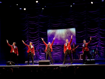 Five men on stage in front of a band behind with a keyboard, guitar, bass, and drum set. The five men are in red glittery suit jackets with black lapels, trousers, and shoes, the two men on the left and right have white shirts and black ties. The centre man is Peter Andre in a black shirt and tie. The two on the left have their right arms up, the two on the right have their left arms up, Peter Andre in the centre has both up.