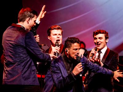 Peter Andre in a purple velvet suit jacket with black lapels and black shirt and tie singing on stage with three other male singers surrounding him who are in the same jacket as Peter but with white shirts with black ties.