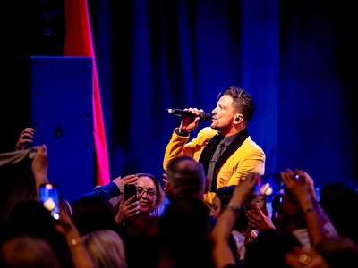 Peter Andre on stage singing in a yellow suit jacket with black lapels and black shirt and tie. There are lots of people in the audience underneath him with their phones up taking photos