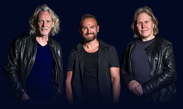 Three members of Wet Wet Wet stood next to each other on a dark background, smiling at the camera