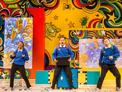 Two men and a woman are dressed in blue school uniform with backpacks on, they all stand in a line across the stage dancing. Behind them are a whole load of doodles on the walls
