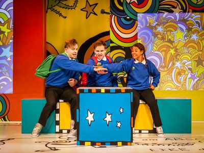 Two men and a woman are dressed in blue school uniform with backpacks on, they all stand around a blue box with white starts on it and put their hands together in the centre above it