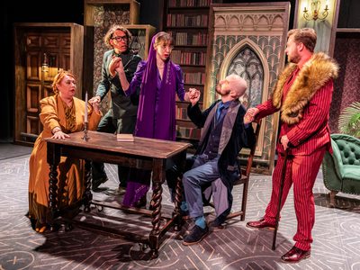 Five people gathered around a table with a candle and book holding hands all looking concerned. From left to right there is a woman in a yellow blouse and skirt with a matching jacket, a man in a green suit with glasses, a woman in purple dress with a long purple scarf draped around her head, a man in a blue suit with tails, and man in a red pin striped suit with a fur shawl and a walking stick. The stage is decorated like an old library with a large stained glass window in the centre.