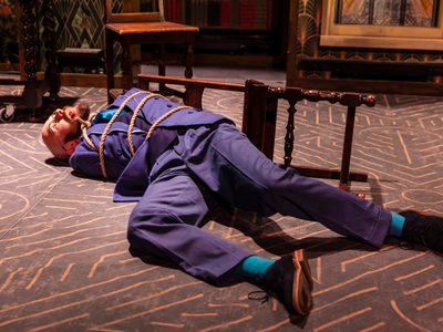 A man dressed in an all blue suit laying on the floor with his tongue sticking out, he is tied to a chair that has fallen over. The stage is decorated like an old library with a large stained glass window in the centre.