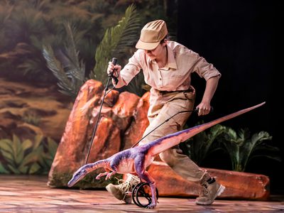 A coelophysis puppet, also with it's legs attached to a wheel, being walked by an actress in a full tan outfit with a button up shirt, trousers, boots, and a cap