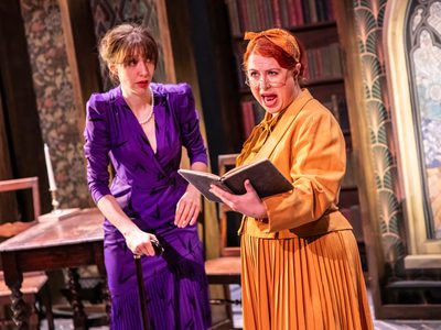 Two women, one dressed in a purple dress with pearls and a walking stick, the other in a yellow shirt, jacket, and skirt with half moon glasses holding a book out in front of her. The woman in purple looks concerned staring at the woman in yellow, while she is shouting. The stage is decorated like an old library with a large stained glass window in the centre.