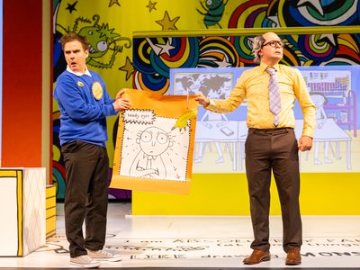 A man dressed in blue school uniform holds up a doodle he's done of his teacher. His teacher, a man with grey hair, glasses and a yellow shirt with a striped tie, looks off into the distance disapprovingly