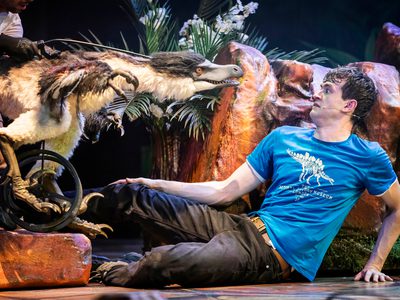 A puppet of a velociraptor, who's legs are attached to a wheel, leering at a scared actor on the floor who's in a blue tshirt