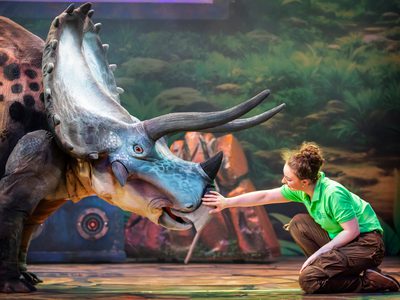 An actress with curly hair in a bun and a green t-shirt kneeling in front of the triceratops puppet petting it