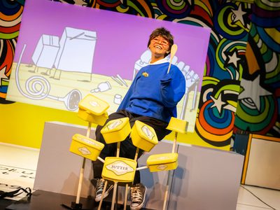 A woman in blue school uniform sits at a drum kit made from butter tubs and wooden spoons