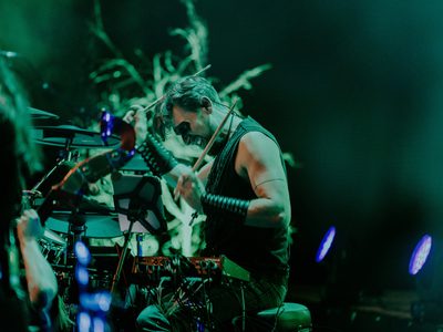 A white man with short hair passionately playing the drums lit in green light. He is wearing a black vest and trousers with large leather bracelets, and has exaggerated black make up around his eyes