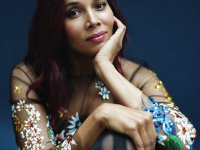 Rhiannon wears a black mesh dress with flowers embroidered on it, she props her elbow on her bent knee and rests her chin in the palm of her hand.