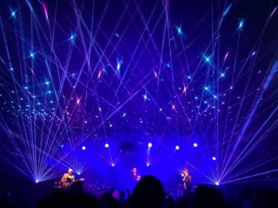 A vibrant concert scene with colourful blue and purple laser lights forming geometric patterns. Three performers are on stage