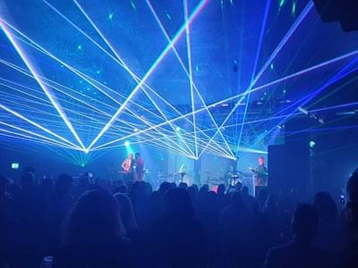 Concert stage with vibrant blue and green laser lights filling the room. Silhouetted crowd watches the performers