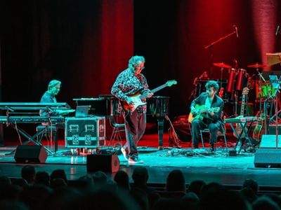 Three men playing instruments on stage under cyan and blue light. They are playing electric and acoustic guitars along with a keyboard