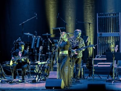 Two men playing acoustic guitar and electric guitars on stage under purple and yellow lighting behind a woman singing