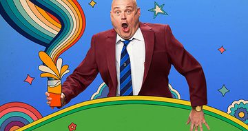 Al Murray with surprised expression, wearing a maroon suit and blue-striped tie, holding a pint with colourful swirls against a vibrant backdrop