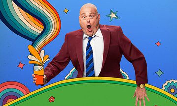 Al Murray with surprised expression, wearing a maroon suit and blue-striped tie, holding a pint with colourful swirls against a vibrant backdrop