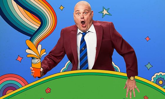 Al Murray with surprised expression, wearing a maroon suit and blue-striped tie, holding a pint with colourful swirls against a vibrant backdrop