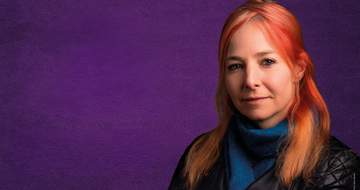 Alice Roberts, a white woman with red hair stands against a purple background. She's wearing a blue turtleneck jumper and a black leather jacket