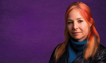 Alice Roberts, a white woman with red hair stands against a purple background. She's wearing a blue turtleneck jumper and a black leather jacket
