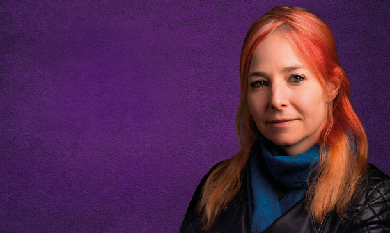 Alice Roberts, a white woman with red hair stands against a purple background. She's wearing a blue turtleneck jumper and a black leather jacket