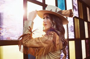 A woman with curled brown hair wears a tan jacket with fringe along the arms and a cream hat with a feather in it
