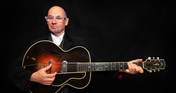 A bald man with clear frame glasses wears a black suit and sits against a black background while holding a guitar