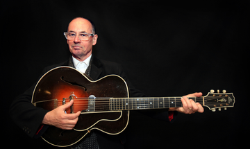 A bald man with clear frame glasses wears a black suit and sits against a black background while holding a guitar