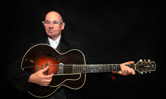 A bald man with clear frame glasses wears a black suit and sits against a black background while holding a guitar