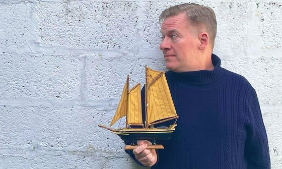 Charlie Connelly in a blue fisherman style jumper holding a model ship and looking to the left