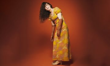 Bridget Christie, a woman in a vibrant yellow and orange floral dress and long brown gloves, posing against an orange background