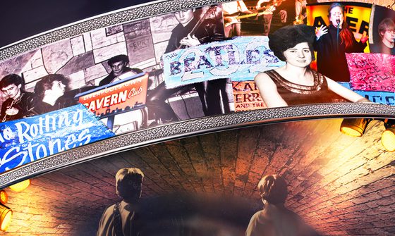 A collage of different artists and their posters that have been to The Cavern Club over the years including The Rolling Stones, The Beatles, Cilla Black, and Adele, on an arch above the brick ceiling of the club with two men looking up at it