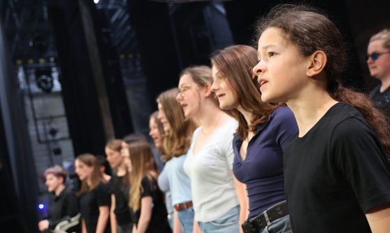 A group of young people in casual clothing stand in rows as they sing together