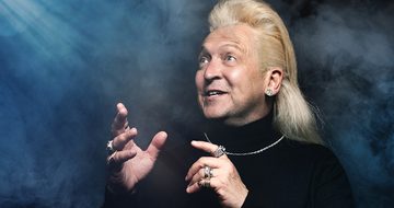 Clinton Baptiste in a black turtle neck with silver chain necklace, various rings on all his fingers, and a large diamond earring. He is looking in awe at a shining light coming trough the dark and fog surrounding him