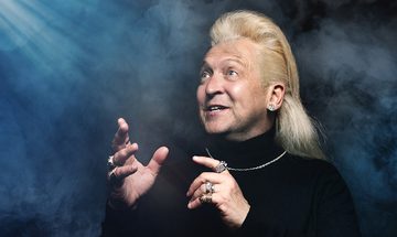 Clinton Baptiste in a black turtle neck with silver chain necklace, various rings on all his fingers, and a large diamond earring. He is looking in awe at a shining light coming trough the dark and fog surrounding him
