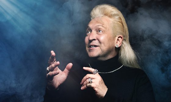 Clinton Baptiste in a black turtle neck with silver chain necklace, various rings on all his fingers, and a large diamond earring. He is looking in awe at a shining light coming trough the dark and fog surrounding him