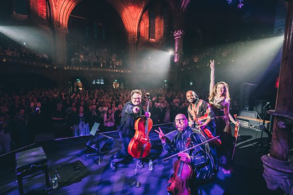 The Vitamin String Quartet, consisting of a bald white man with a beard and glasses, a white man with medium length brown hair, a moustache, and glasses, a bald black man with a beard, and a white woman with long blonde hair all posing and smiling to the camera on stage holding their instruments with the audience behind them