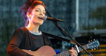 Edwina wears a black dress with a bejewelled collar, she sings into a microphone while playing the acoustic guitar