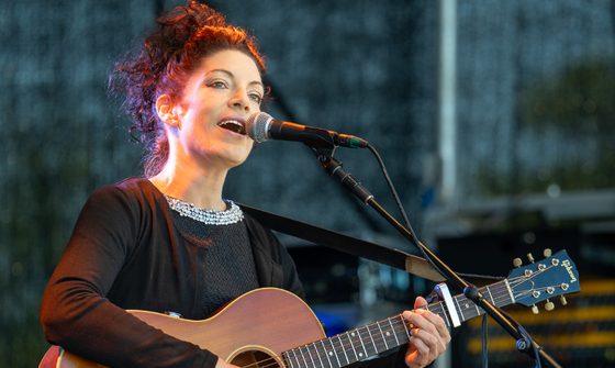 Edwina wears a black dress with a bejewelled collar, she sings into a microphone while playing the acoustic guitar