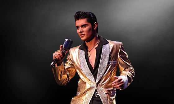 Emilio in a black shirt and gold blazer, with his black hair quaffed like Elvis' holding a microphone